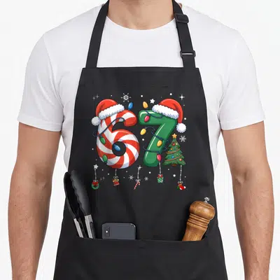 Meme six christmas apron - Number 67 wearing Santa hats wrapped in lights with ornaments and a tree — shown on a black cotton canvas apron against a white background