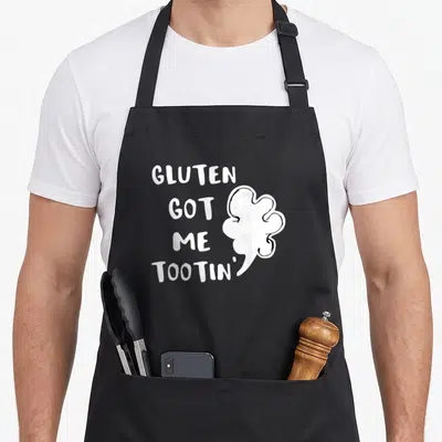 Gluten celiac awareness apron - White stacked text reading GLUTEN GOT ME TOOTIN' beside a cartoon gas cloud — shown on a black cotton canvas apron against a white background