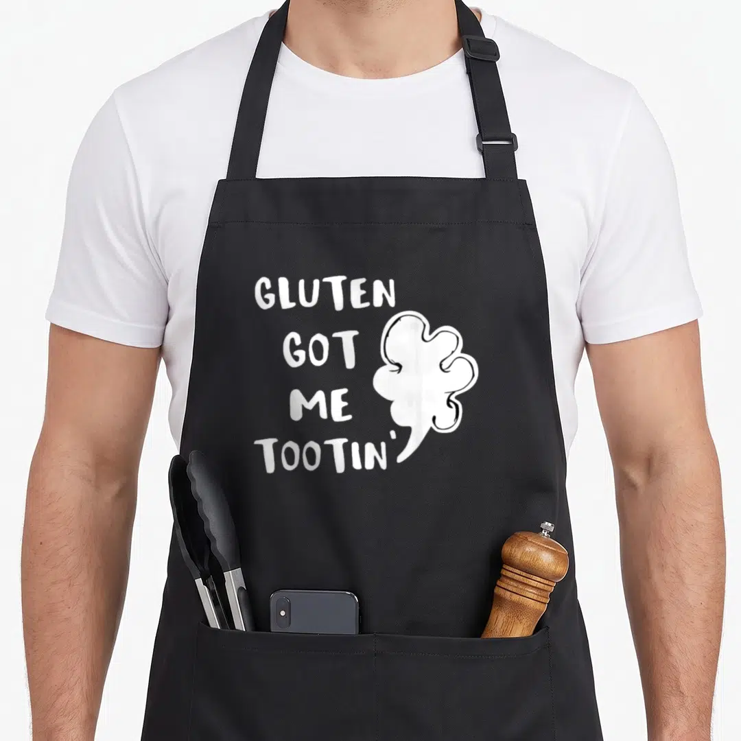 Gluten celiac awareness apron - White stacked text reading GLUTEN GOT ME TOOTIN' beside a cartoon gas cloud — shown on a black cotton canvas apron against a white background