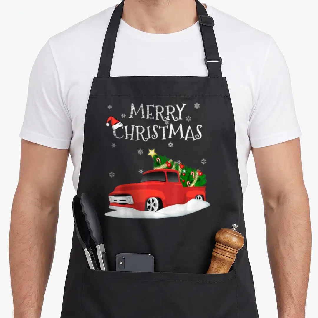 Truck pickup christmas tree apron - Red truck carrying decorated tree under stacked Merry Christmas text with Santa hat — shown on a black cotton canvas apron against a white background