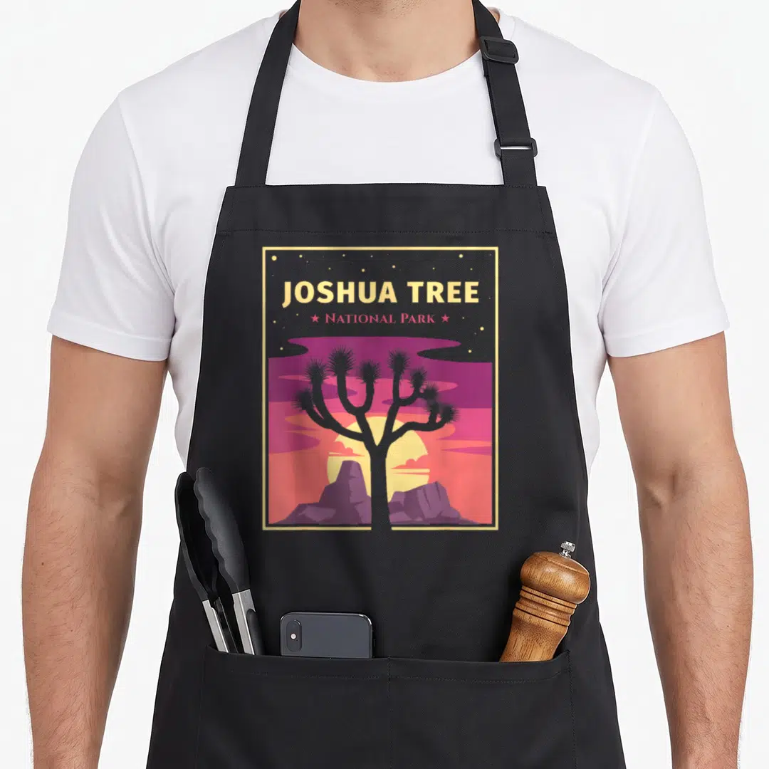 Joshua national park tree apron - Retro poster style joshua tree silhouette against purple and pink sunset with stacked — shown on a black cotton canvas apron against a white background