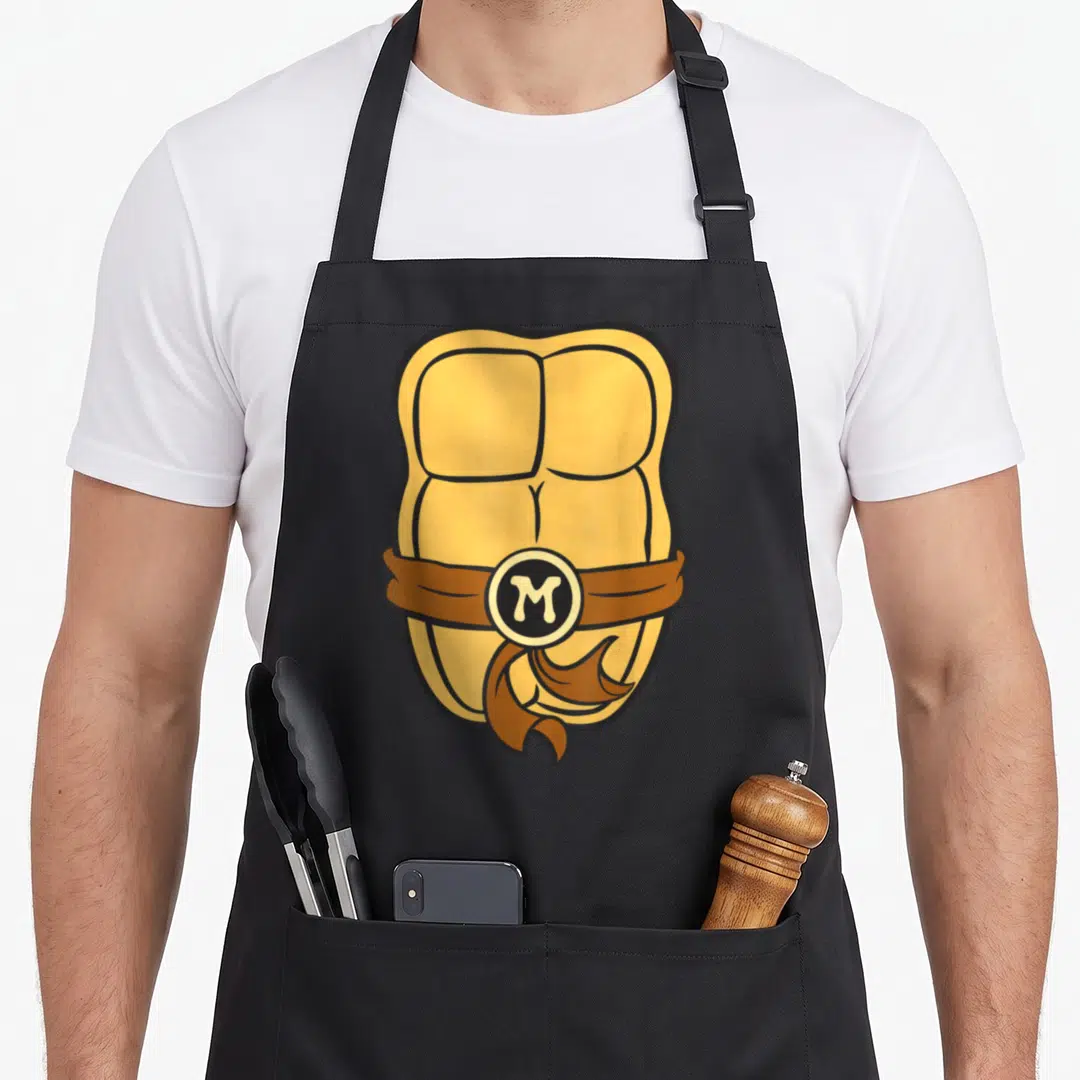 Michaelangelo teenage mutant ninja apron - Cartoon yellow abs with brown belt and circular M buckle — shown on a black cotton canvas apron against a white background