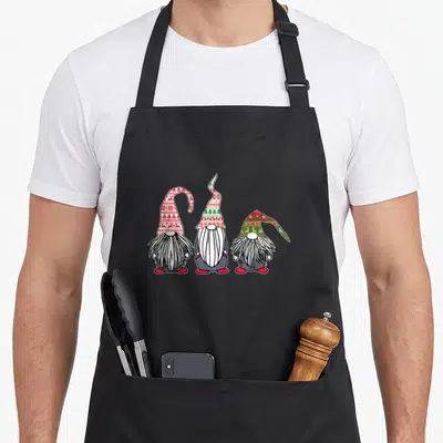 Nordic swedish apron - Three cartoon gnomes with red white green grey patterned hats standing side by side — shown on a black cotton canvas apron against a white background