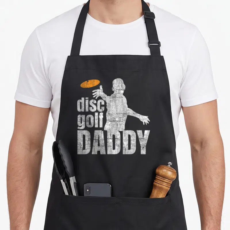 Disc daddy apron - Distressed white golfer silhouette throwing orange disc with stacked disc golf DADDY text — shown on a black cotton canvas apron against a white background