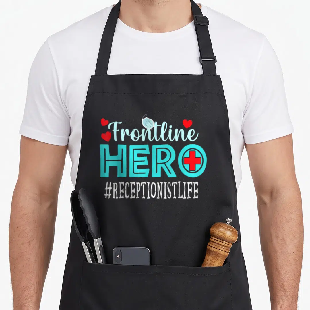 Receptionist essential apron - Stacked Frontline Hero text with face mask, red cross, and hearts — shown on a black cotton canvas apron against a white background