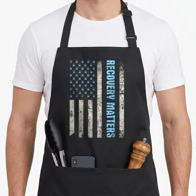 Recovery weird being apron - Distressed flag with light blue ribbon grid and vertical RECOVERY MATTERS text on black — shown on a black cotton canvas apron against a white background