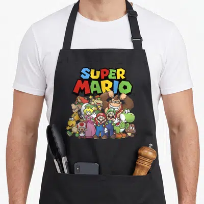 Cast shot apron - SUPER MARIO text above Mario Luigi Peach Yoshi Donkey Kong Bowser Toad Goomba yellow creature — shown on a black cotton canvas apron against a white background