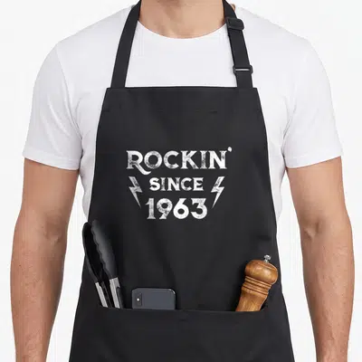 Rock 63rd birthday apron - White distressed text ROCKIN SINCE 1963 with two lightning bolts on black fabric — shown on a black cotton canvas apron against a white background