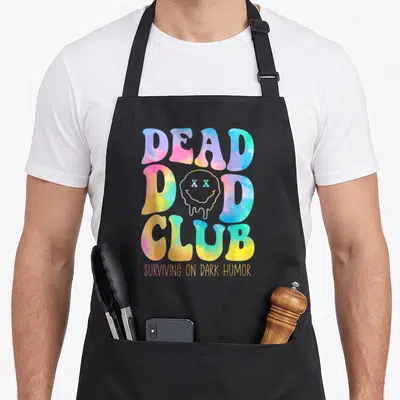 Dead hippie apron - DEAD DAD CLUB bubble text with melting smiley face and dark humor caption in tie-dye colors — shown on a black cotton canvas apron against a white background