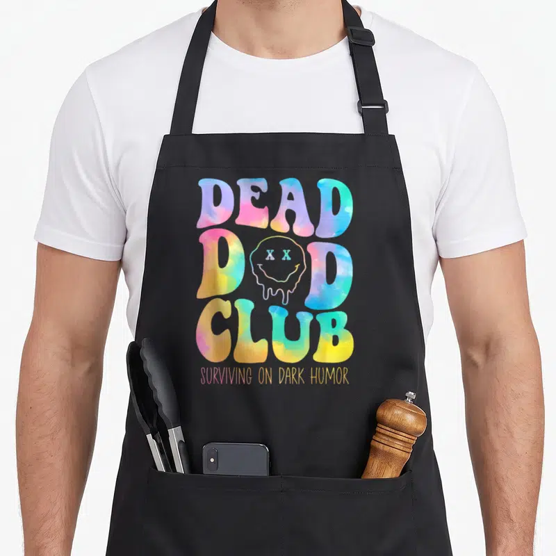 Dead hippie apron - DEAD DAD CLUB bubble text with melting smiley face and dark humor caption in tie-dye colors — shown on a black cotton canvas apron against a white background