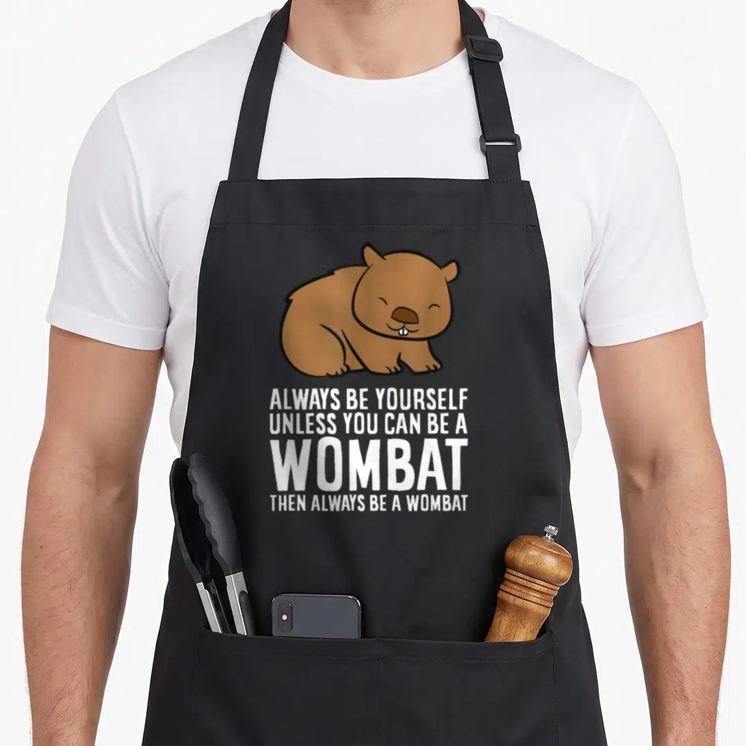 Wombat yourself unless apron - Brown cartoon wombat above stacked white text reading Always Be Yourself Unless You Can — shown on a black cotton canvas apron against a white background