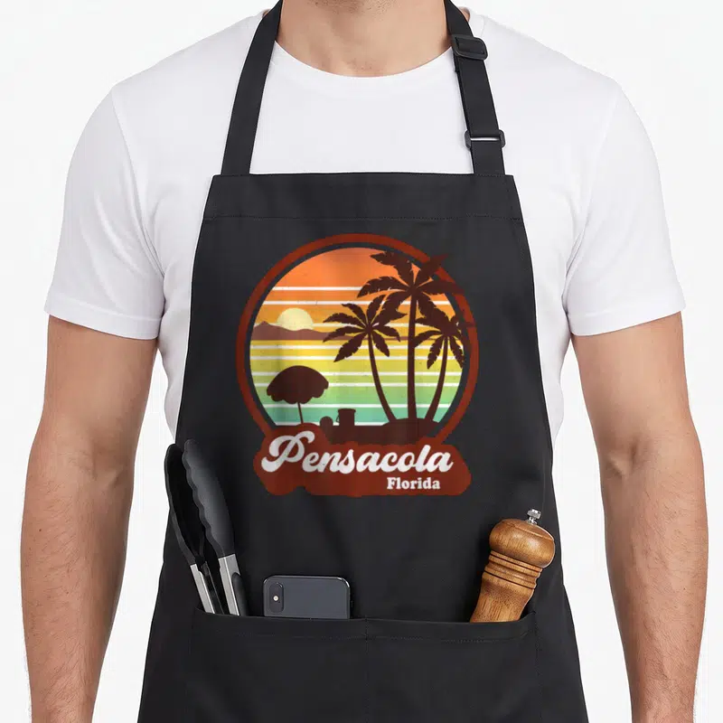 Pensacola summer aprons - Retro circular sunset with palm trees and Pensacola Florida text in brown and orange — shown on a black cotton canvas apron against a white background