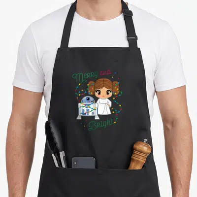 Bright merry christmas apron - Cartoon girl and robot droid wrapped in colorful lights with Merry and Bright text — shown on a black cotton canvas apron against a white background