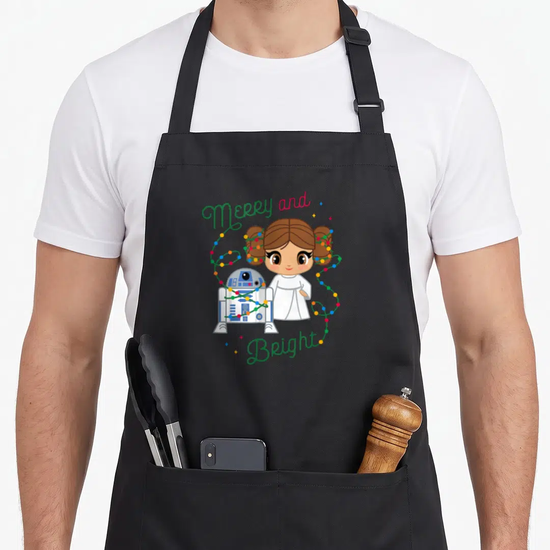 Bright merry christmas apron - Cartoon girl and robot droid wrapped in colorful lights with Merry and Bright text — shown on a black cotton canvas apron against a white background