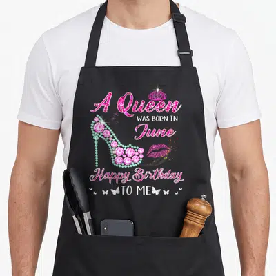 June birthday baking queen apron - Pink jeweled high heel shoe, crown, lips, and stacked birthday text reading A Queen — shown on a black cotton canvas apron against a white background