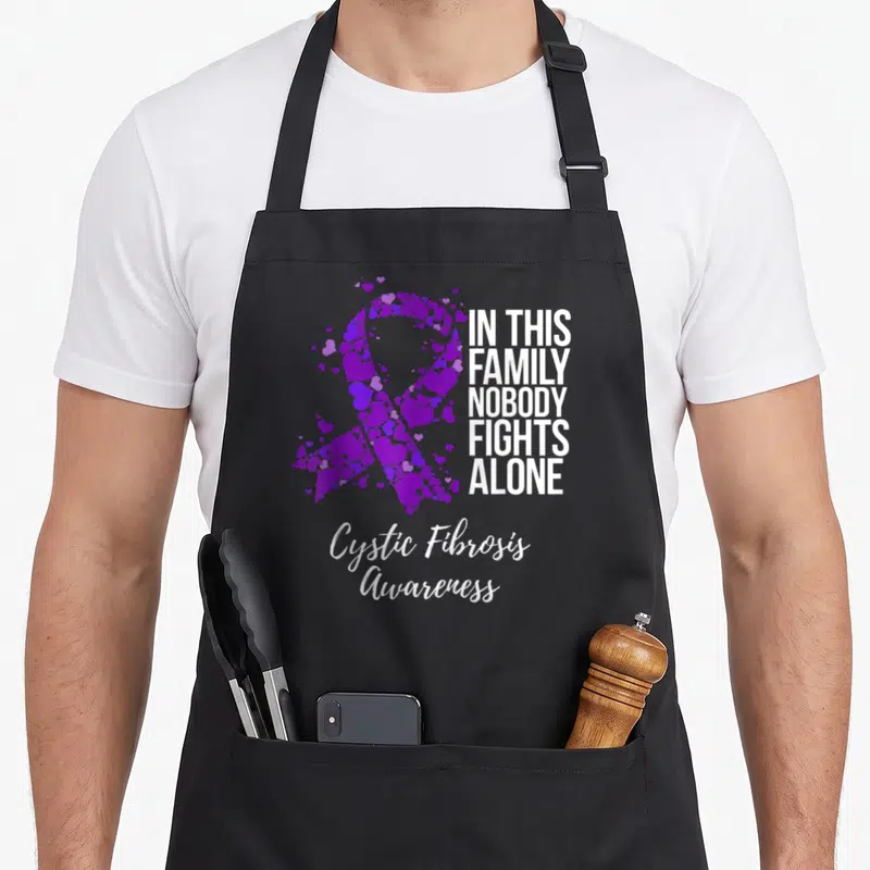 Cystic family aprons - Purple ribbon formed by hearts next to white stacked text reading In This Family Nobody Fights — shown on a black cotton canvas apron against a white background