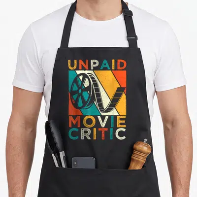 Unpaid fan apron - Retro film reel on orange, yellow, teal, grey stripes with UNPAID MOVIE CRITIC text — shown on a black cotton canvas apron against a white background