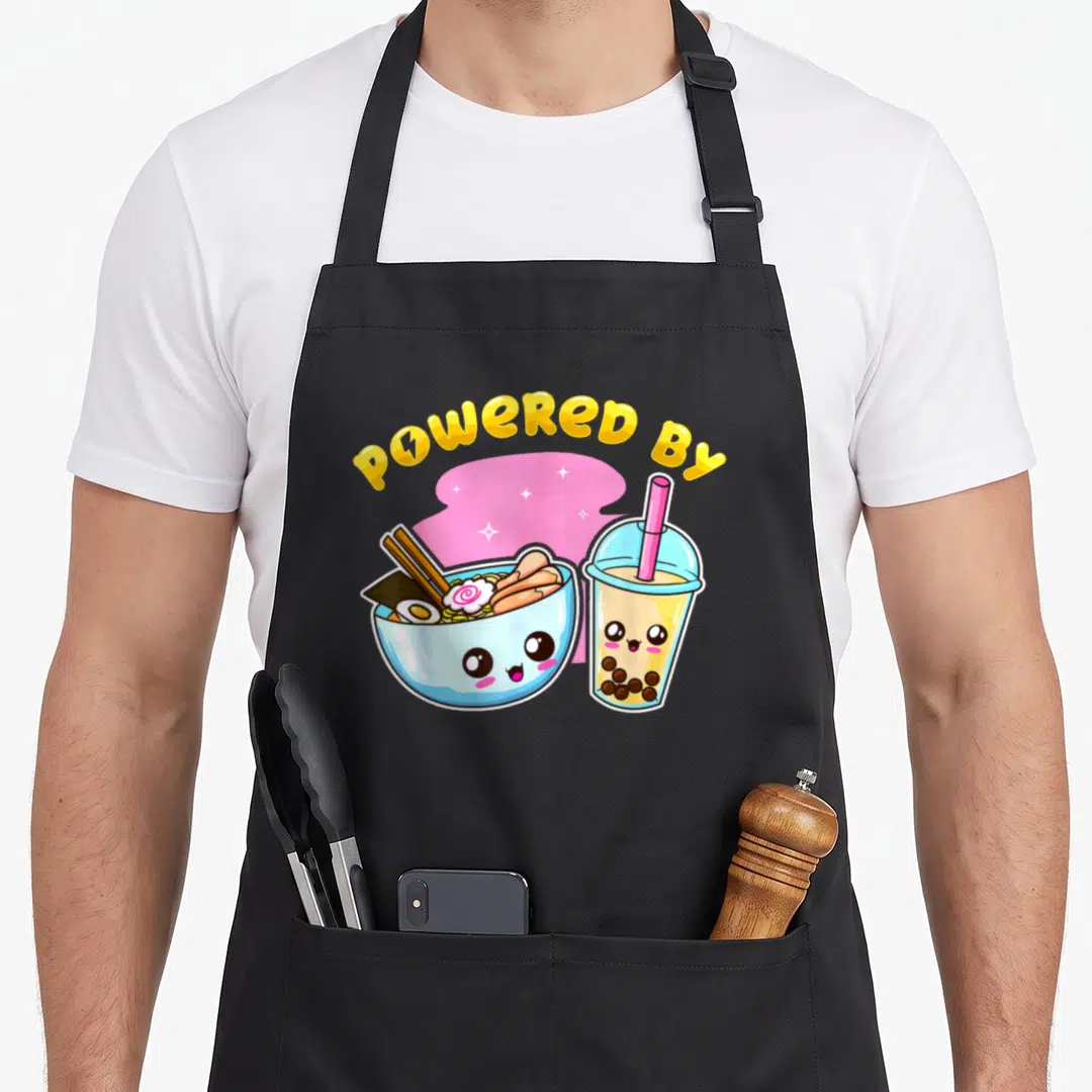 Bubble anime apron - POWERED BY text over cartoon ramen bowl and boba tea cup with pink stars — shown on a black cotton canvas apron against a white background