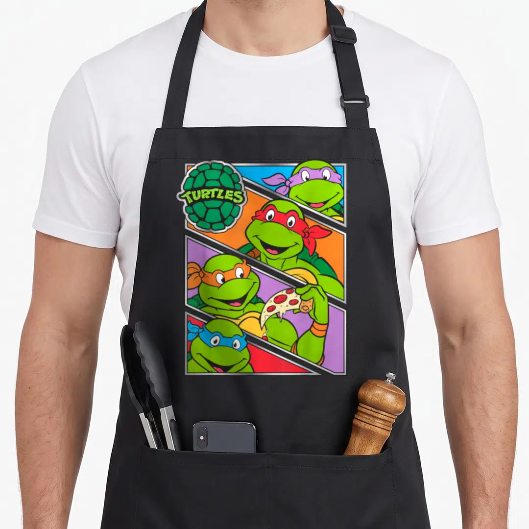 Multiple teenage cooking aprons - Four cartoon turtles in diagonal panels, one holding pizza, TURTLES shell logo — shown on a black cotton canvas apron against a white background