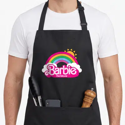 Movie rainbow apron - Barbie The Movie logo under rainbow arch with sun and white clouds in pink yellow blue — shown on a black cotton canvas apron against a white background