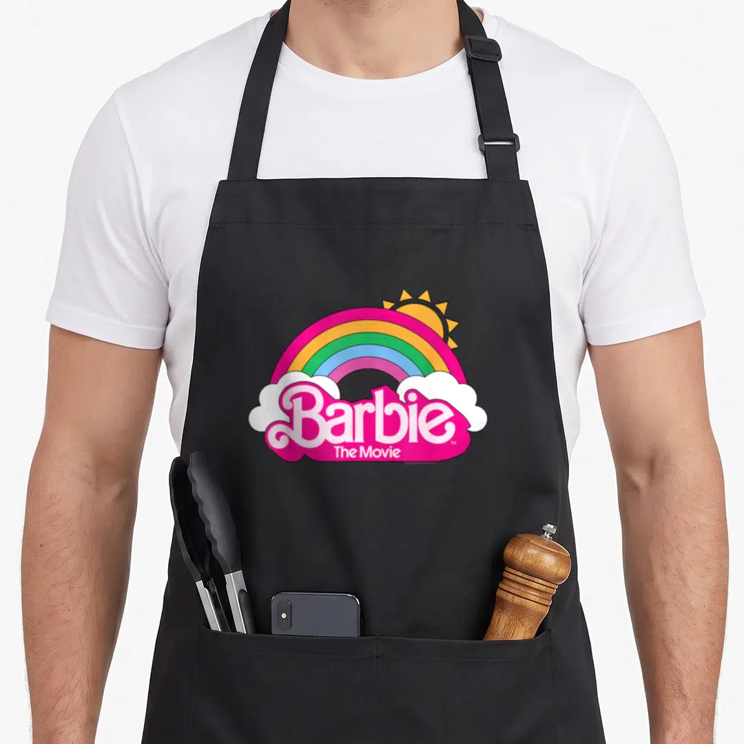 Movie rainbow apron - Barbie The Movie logo under rainbow arch with sun and white clouds in pink yellow blue — shown on a black cotton canvas apron against a white background