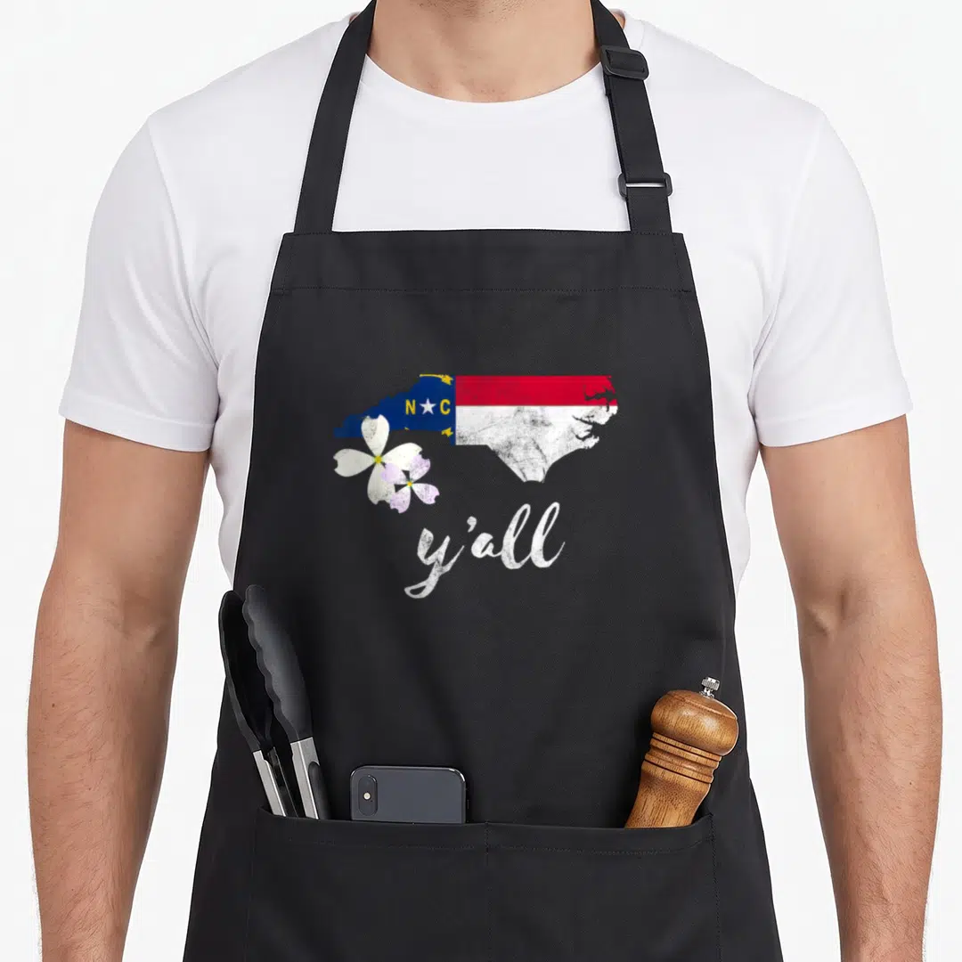 State dogwood apron - North Carolina map in flag colors with two dogwood flowers and y'all text below — shown on a black cotton canvas apron against a white background
