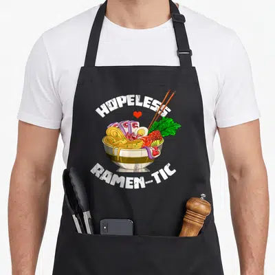 Noodle valentine's day apron - Cartoon ramen bowl with HOPELESS RAMEN-TIC text, red heart, and chopsticks — shown on a black cotton canvas apron against a white background