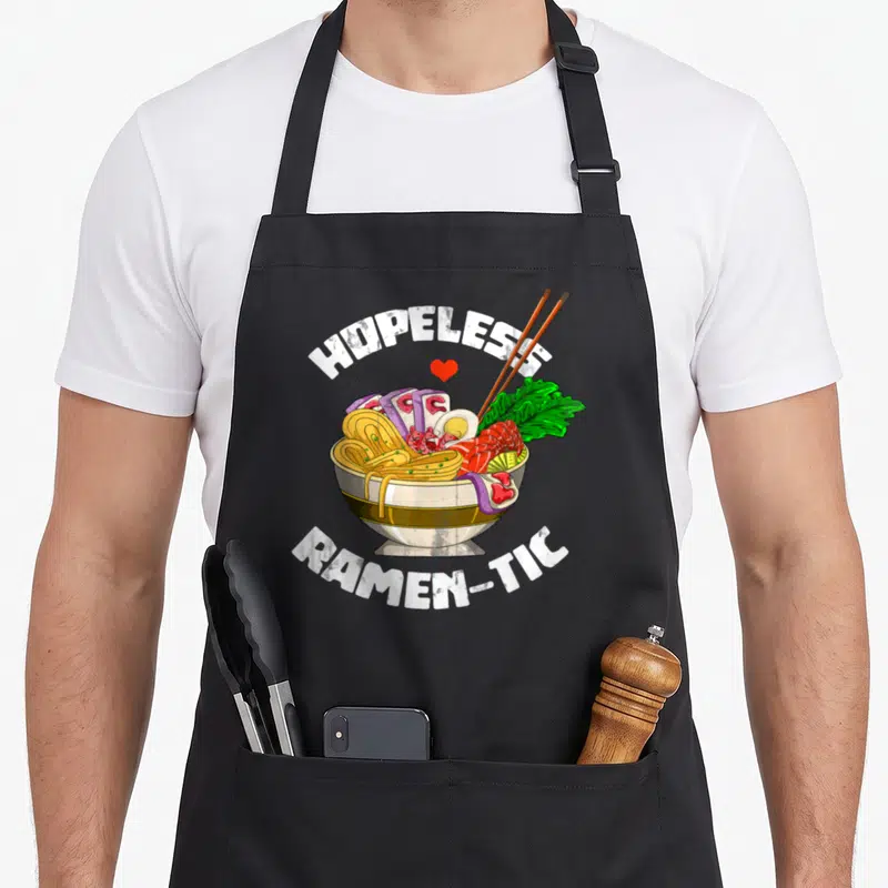 Noodle valentine's day apron - Cartoon ramen bowl with HOPELESS RAMEN-TIC text, red heart, and chopsticks — shown on a black cotton canvas apron against a white background