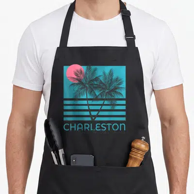 Charleston souvenir tree apron - Retro graphic of two black palm trees and pink sun on blue background with grey — shown on a black cotton canvas apron against a white background