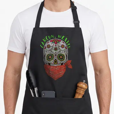 Cancun sugar skull apron - Cartoon sugar skull with red bandana and green CANCUN MEXICO text — shown on a black cotton canvas apron against a white background