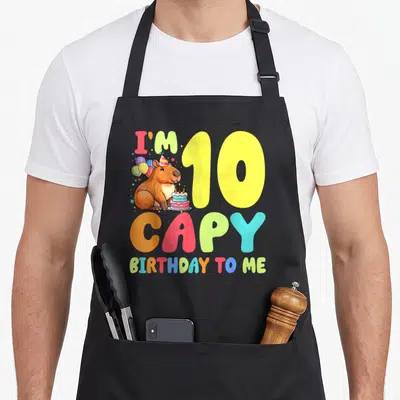 Capy 10th birthday apron - Capybara wearing party hat with balloons and cake next to colorful I'm 10 CAPY text — shown on a black cotton canvas apron against a white background