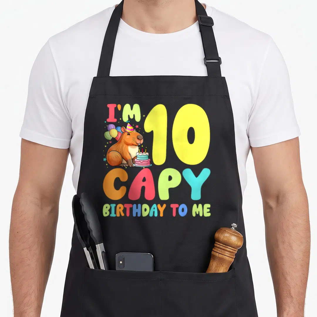 Capy 10th birthday apron - Capybara wearing party hat with balloons and cake next to colorful I'm 10 CAPY text — shown on a black cotton canvas apron against a white background