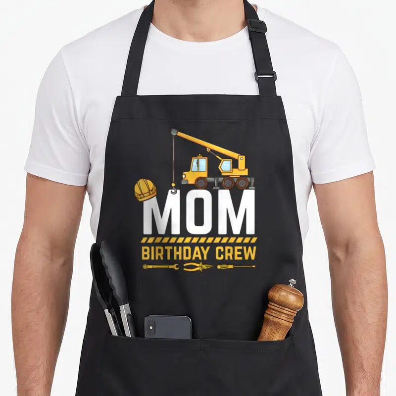 Birthday construction crew aprons - Cartoon crane lifting white MOM text above yellow BIRTHDAY CREW and tools — shown on a black cotton canvas apron against a white background