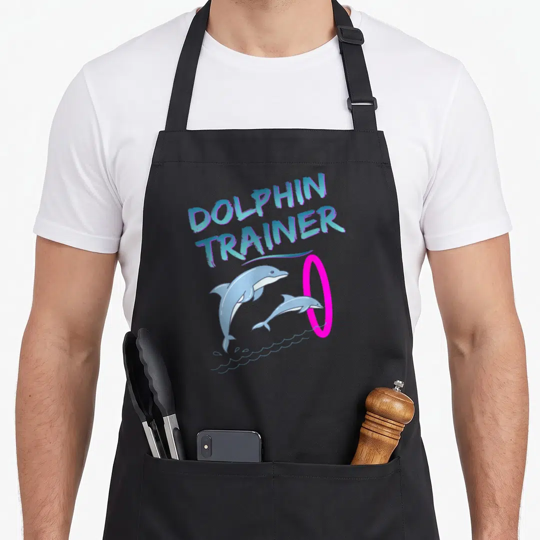 Trainer halloween apron - Two cartoon dolphins jumping through a pink hoop under arched DOLPHIN TRAINER text — shown on a black cotton canvas apron against a white background