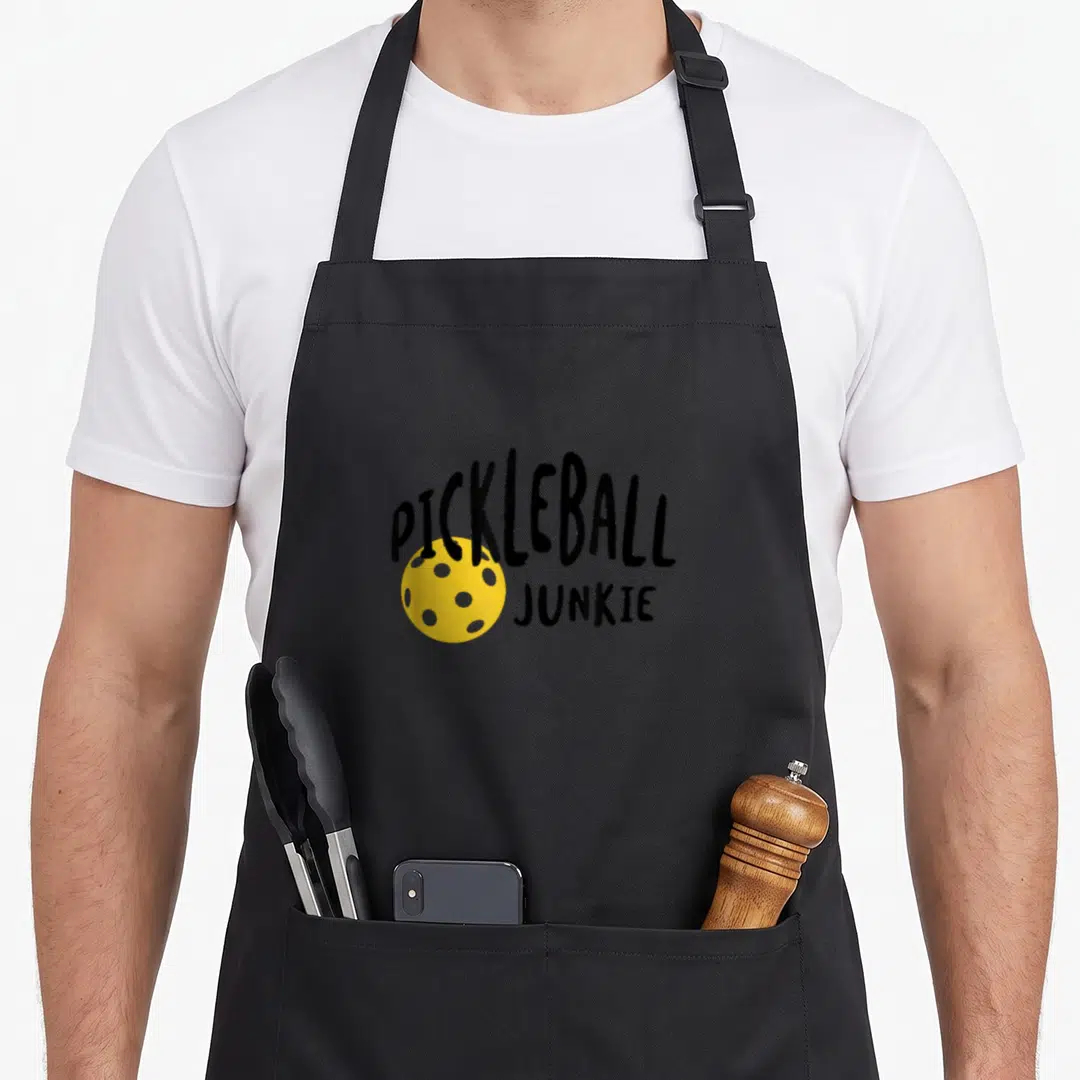 Junkie pickleball apron - Black text PICKLEBALL JUNKIE arched over a yellow dotted ball — shown on a black cotton canvas apron against a white background
