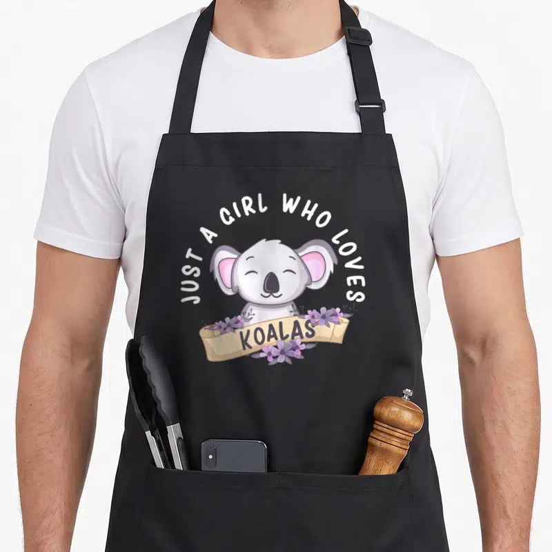 Koalas bear apron - Cartoon koala with JUST A GIRL WHO LOVES KOALAS text and purple flowers on beige ribbon — shown on a black cotton canvas apron against a white background
