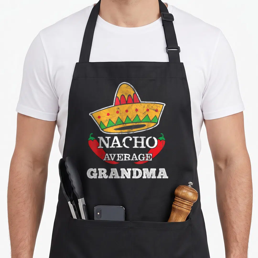 Nacho grandma apron - Yellow sombrero with red dots and green zigzags above two red chilies framing white NACHO AVERAGE — shown on a black cotton canvas apron against a white background