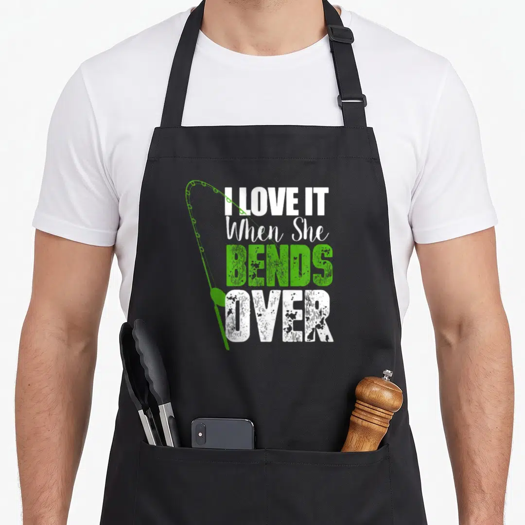 She fishing aprons - I LOVE IT When She BENDS OVER text with green fishing rod curving left over four stacked lines — shown on a black cotton canvas apron against a white background