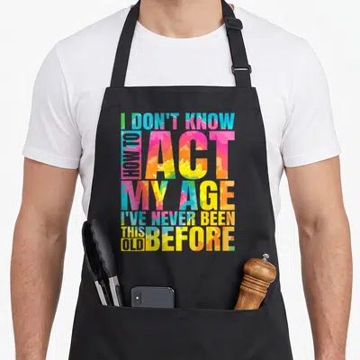 Retro act age apron - Colorful stacked text I DON'T KNOW HOW TO ACT MY AGE I'VE NEVER BEEN THIS OLD BEFORE — shown on a black cotton canvas apron against a white background