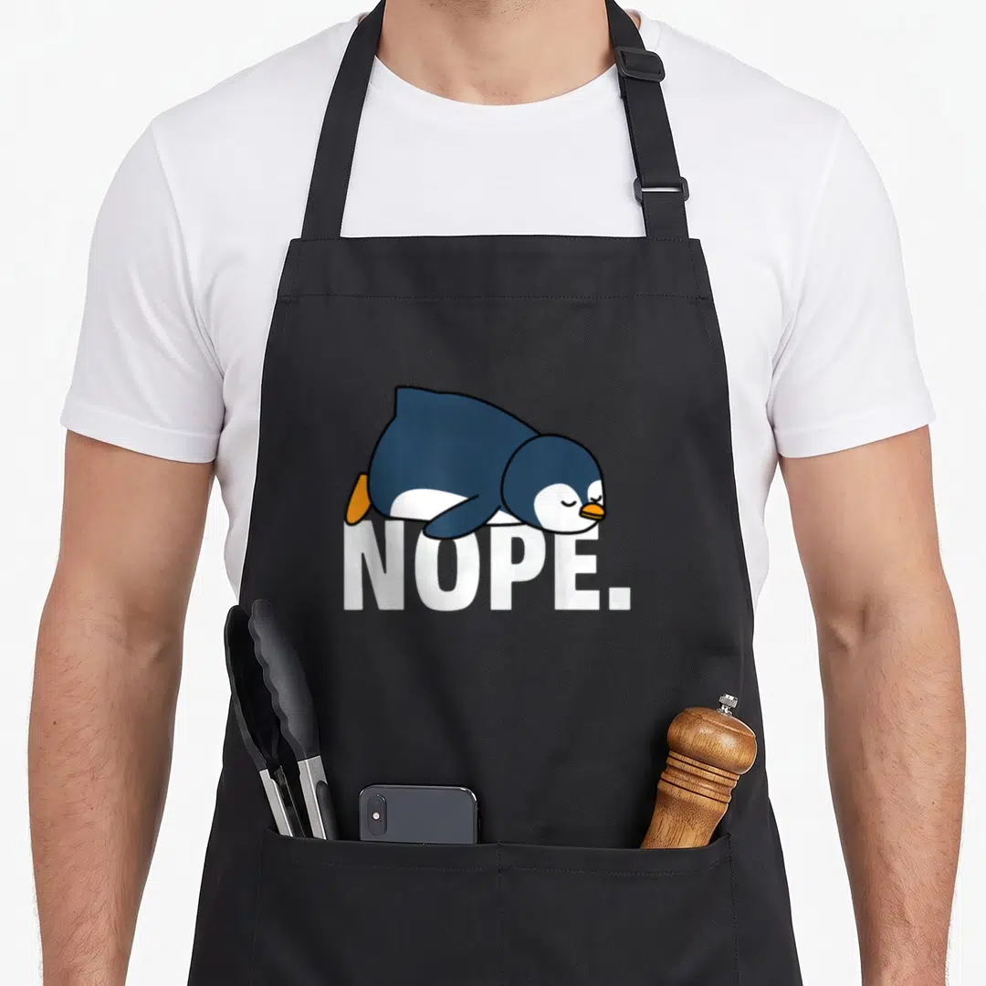 Nope penguin aprons - Cartoon blue penguin lying flat on white NOPE text — shown on a black cotton canvas apron against a white background
