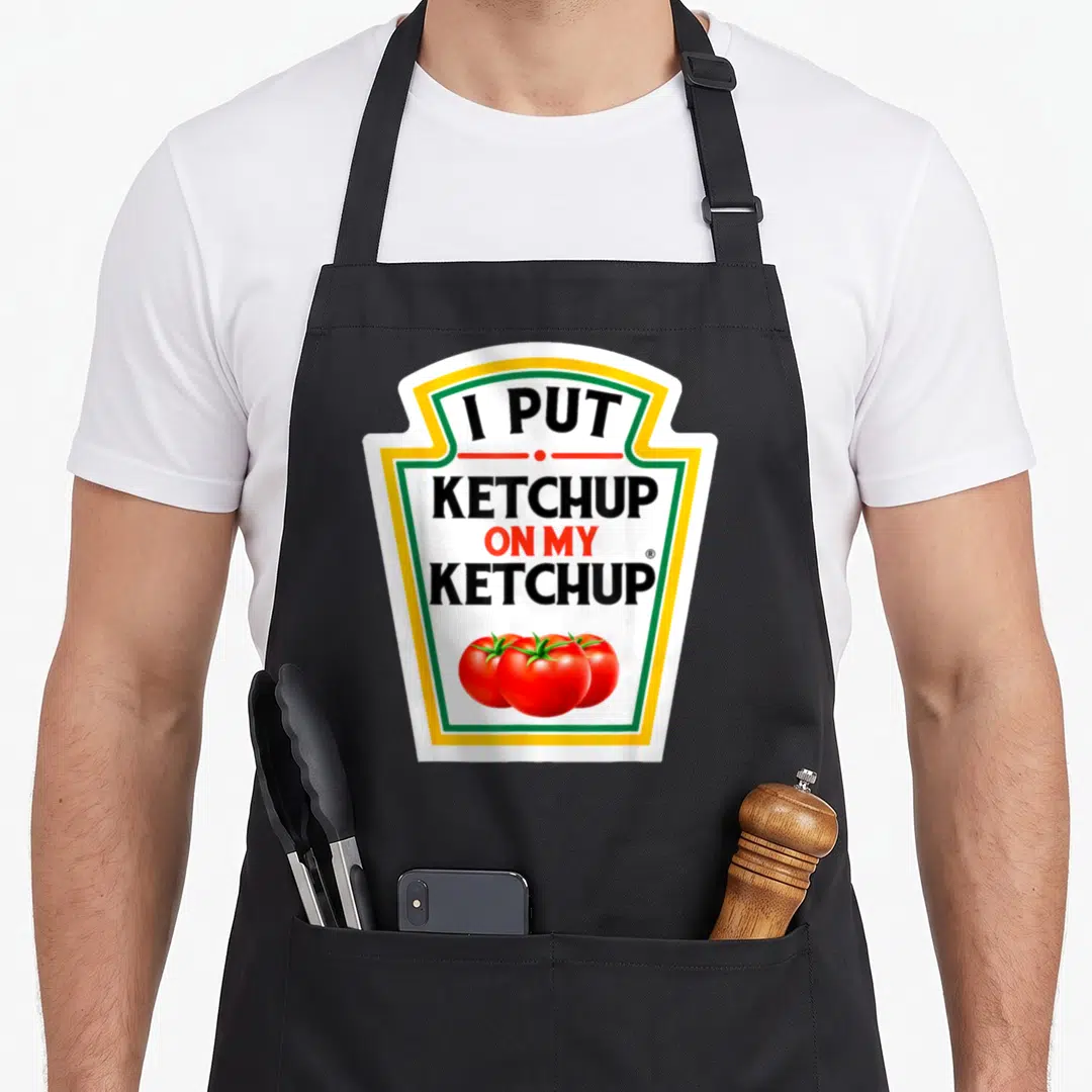 Ketchup tomato halloween apron - Shield label reading I PUT KETCHUP ON MY KETCHUP with three red tomatoes at the bottom — shown on a black cotton canvas apron against a white background