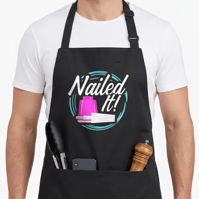 Nailed manicurist apron - Pink nail polish bottle and brush with Nailed It! text in light blue circle — shown on a black cotton canvas apron against a white background