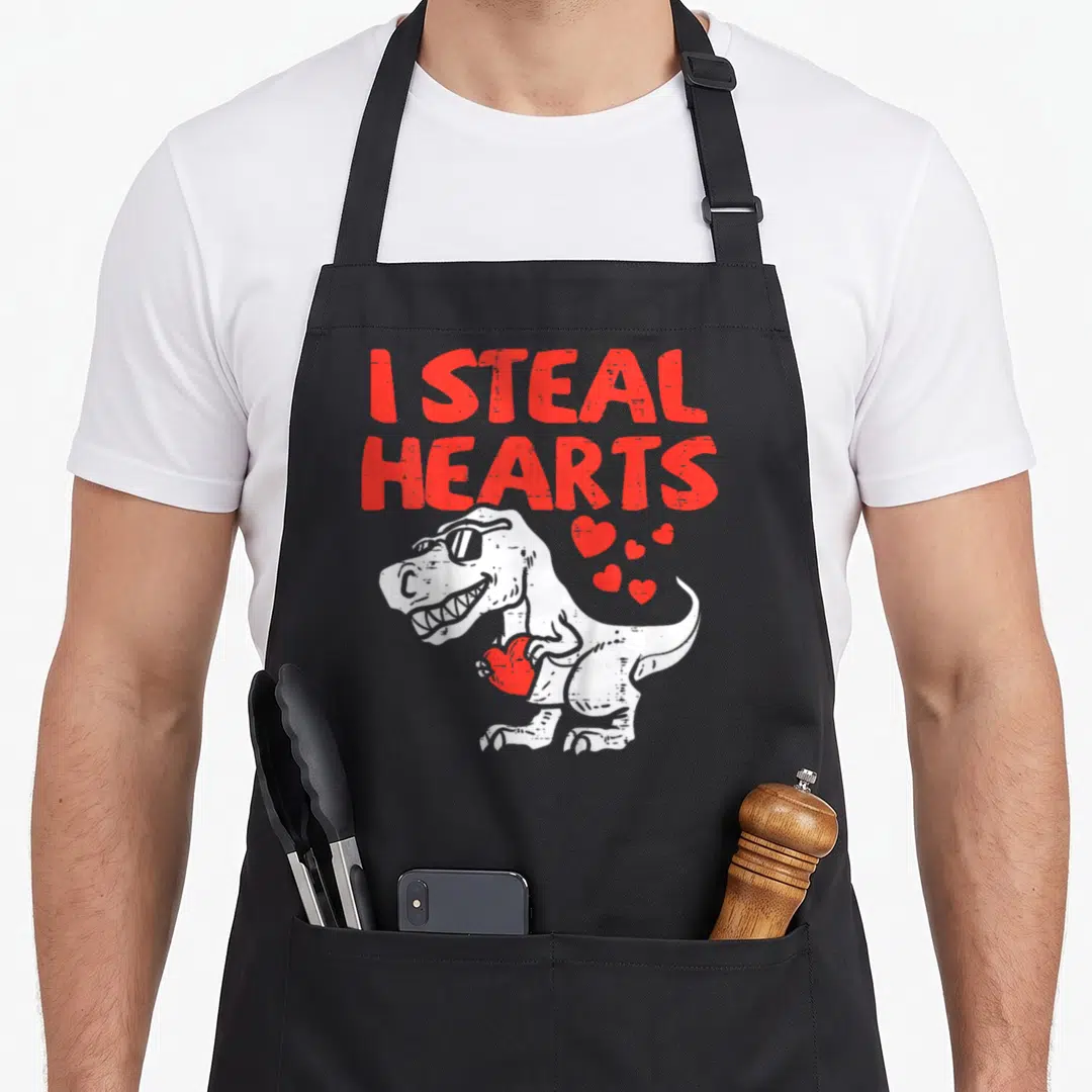 Steal valentine apron - White T-Rex wearing sunglasses holding a red heart below red I STEAL HEARTS text — shown on a black cotton canvas apron against a white background