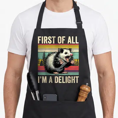 Delight opossum apron - Retro graphic of snarling opossum over red yellow green stripes with text FIRST OF ALL I'M — shown on a black cotton canvas apron against a white background