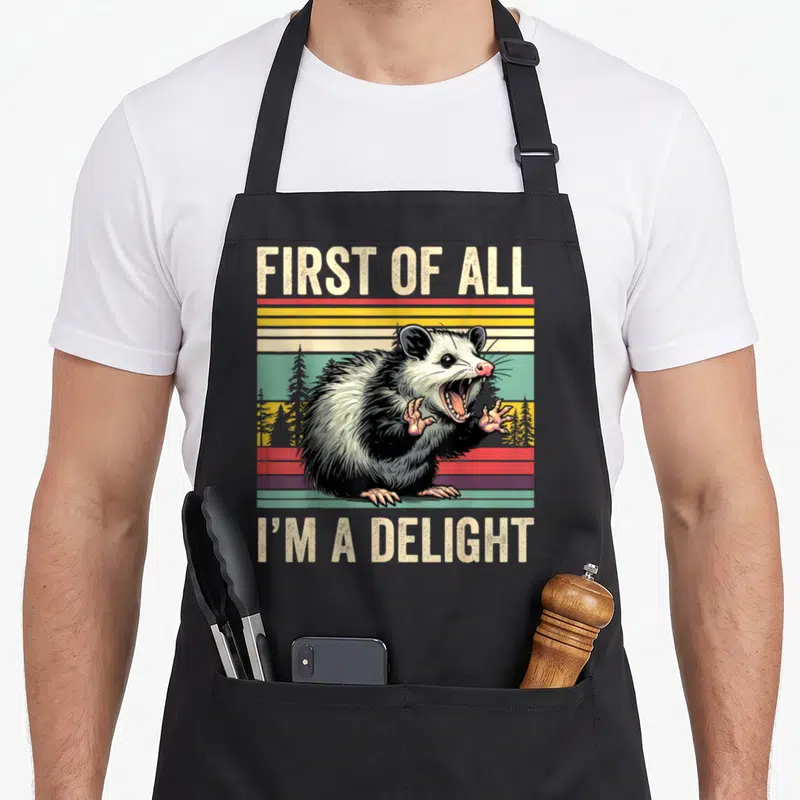 Delight opossum apron - Retro graphic of snarling opossum over red yellow green stripes with text FIRST OF ALL I'M — shown on a black cotton canvas apron against a white background