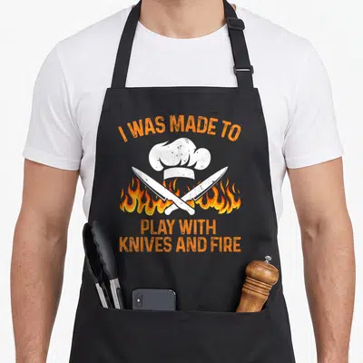 Vintage camping culinary aprons - Chef hat and two crossed knives over orange flames with text I WAS MADE TO PLAY — shown on a black cotton canvas apron against a white background