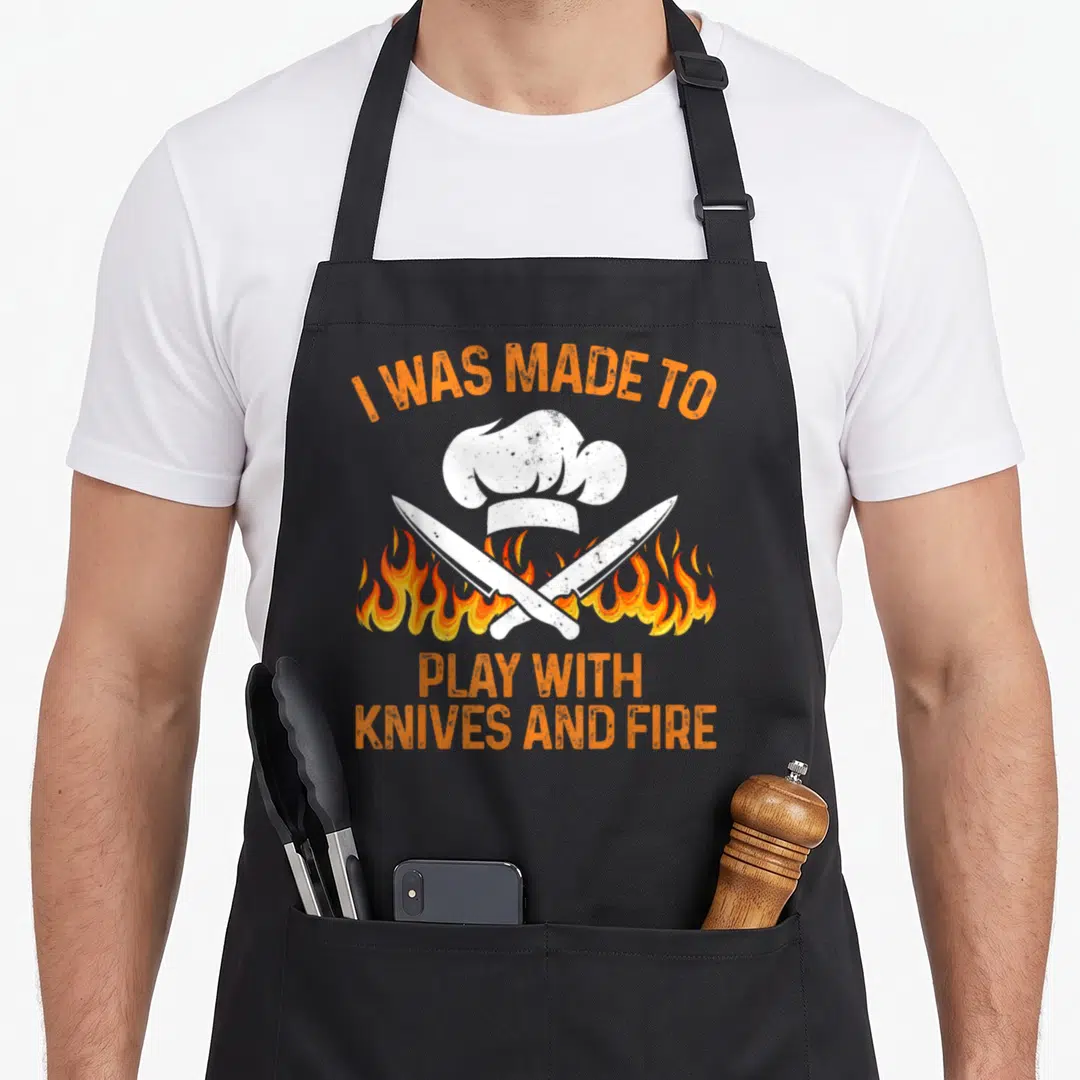 Vintage camping culinary aprons - Chef hat and two crossed knives over orange flames with text I WAS MADE TO PLAY — shown on a black cotton canvas apron against a white background