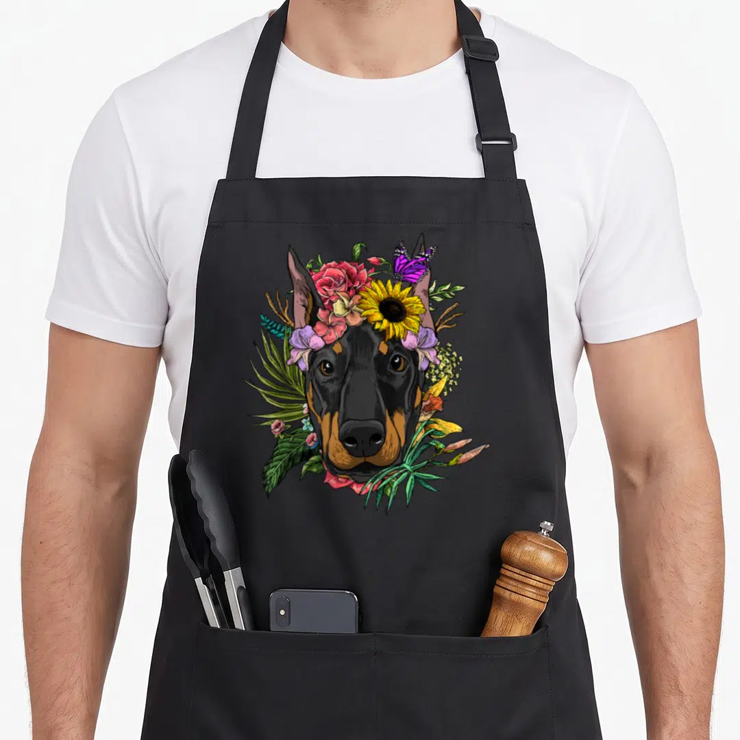 Pinscher floral apron - Cartoon doberman head surrounded by pink, yellow, purple flowers and butterfly on black — shown on a black cotton canvas apron against a white background