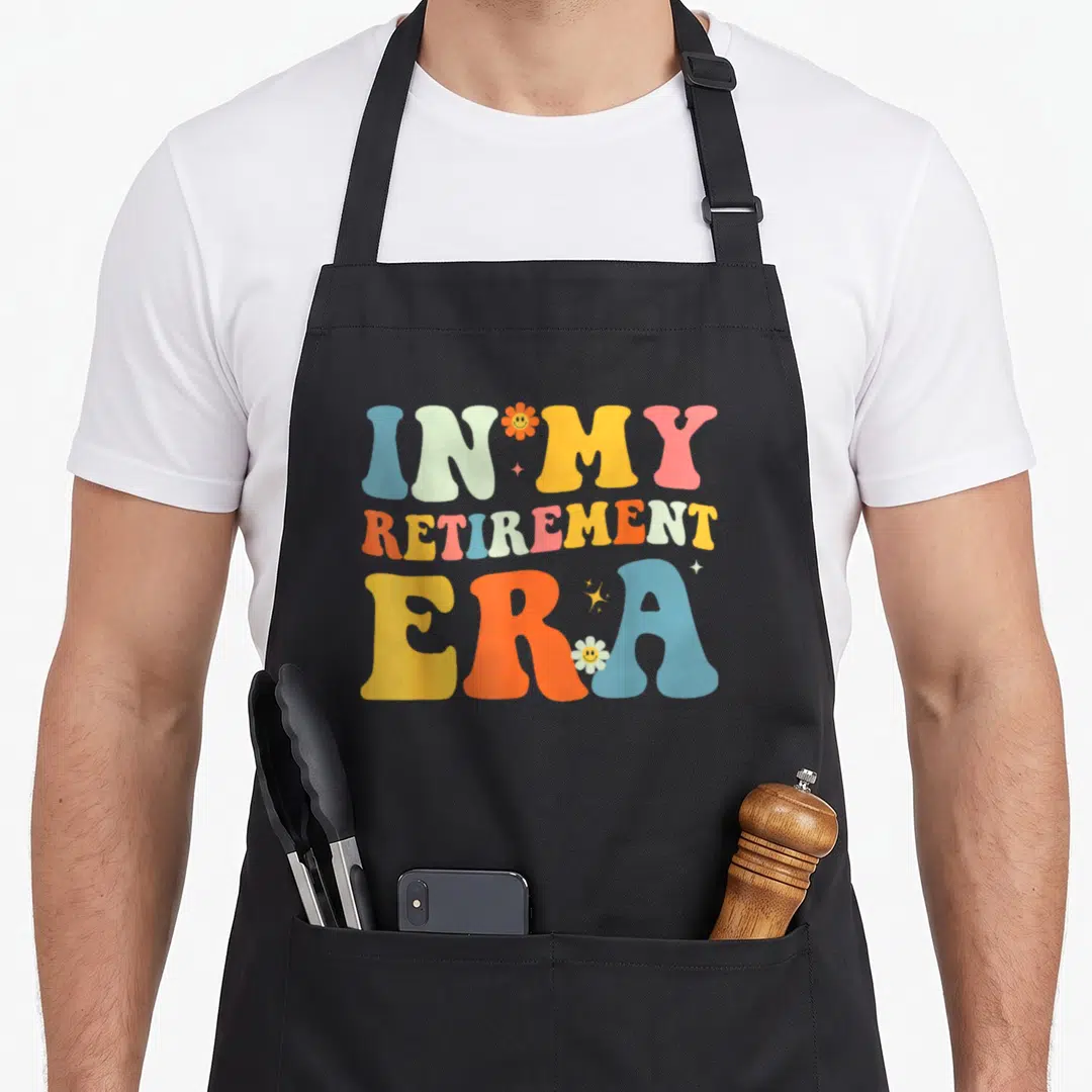 Era retirement apron - Stacked retro text IN MY RETIREMENT ERA with smiley flowers in blue yellow orange pink light — shown on a black cotton canvas apron against a white background
