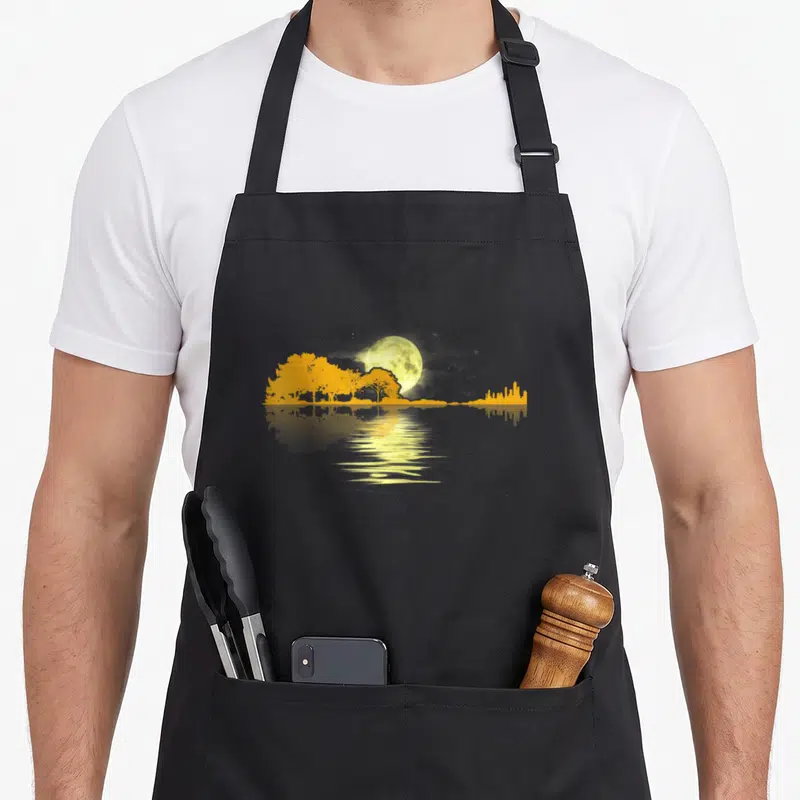 Guitar musician apron - Yellow moon and grey skyline silhouette reflected in water on black fabric — shown on a black cotton canvas apron against a white background